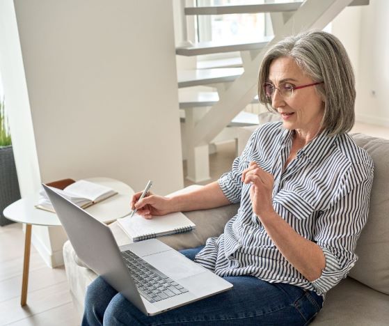 harmaahiuksinen nainen istuu sohvalla, katsoo sylissä olevaa tietokonetta ja kirjoittaa vihkoon