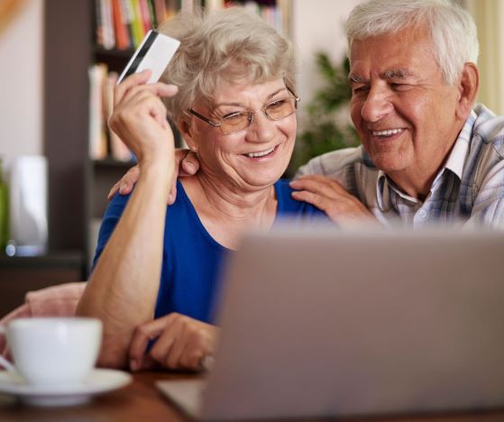 Ikääntyneet nainen ja mies katsovat iloisena tietokonetta ja naisella on luottokortti kädessä